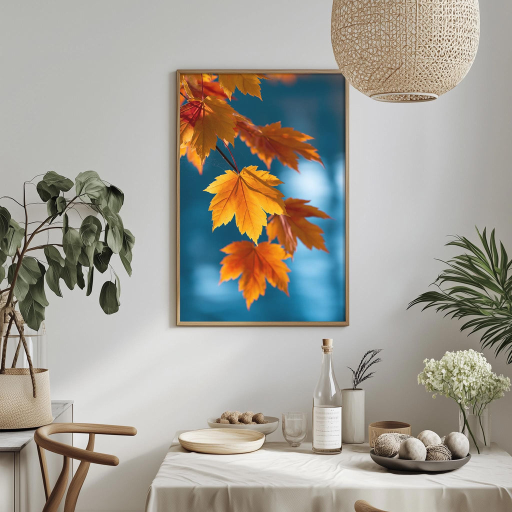 a dining table with a white tablecloth, a bowl of fruit, a bottle, and a vase with flowers. Above the table, there is a framed picture of orange and yellow autumn leaves.
