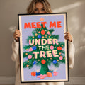 A woman is holding up a framed poster with a Christmas tree design and the text "MEET ME UNDER THE TREE" written on it.