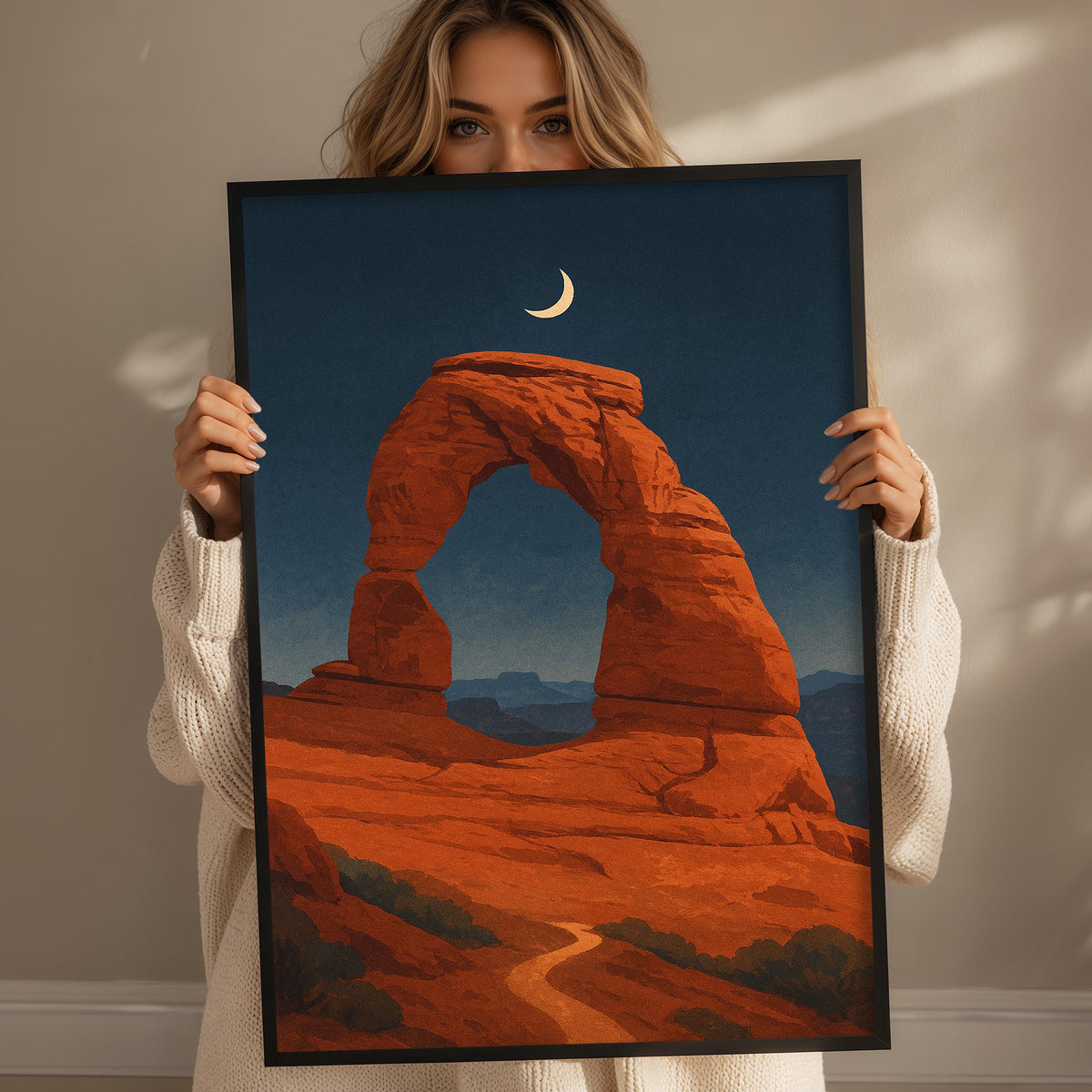 A woman holds up a framed print of a desert landscape with a crescent moon in the sky.