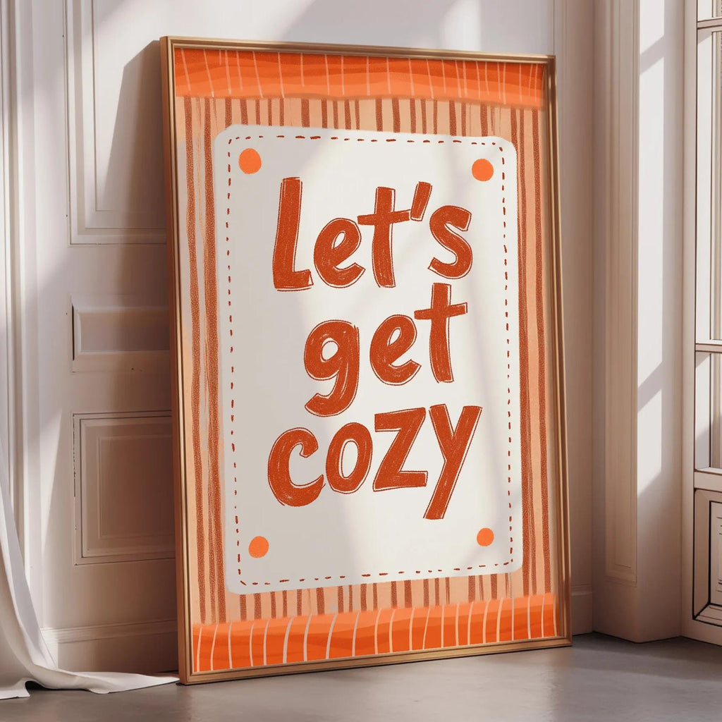 A framed poster with the text "Let's get cozy" displayed prominently on it, placed on a wooden floor next to a window.