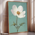 A framed painting of a white flower with a green stem and leaves is displayed on a wall in a room with a window.