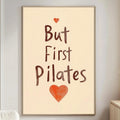 A living room with a white sofa, a wooden side table, and a framed poster with the text "But First Pilates" and an orange heart symbol.