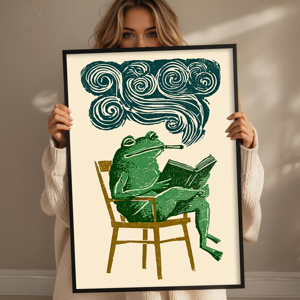 A woman is holding up a framed print that depicts a green frog sitting in a chair and reading a book, with a cloud-like design above the frog.