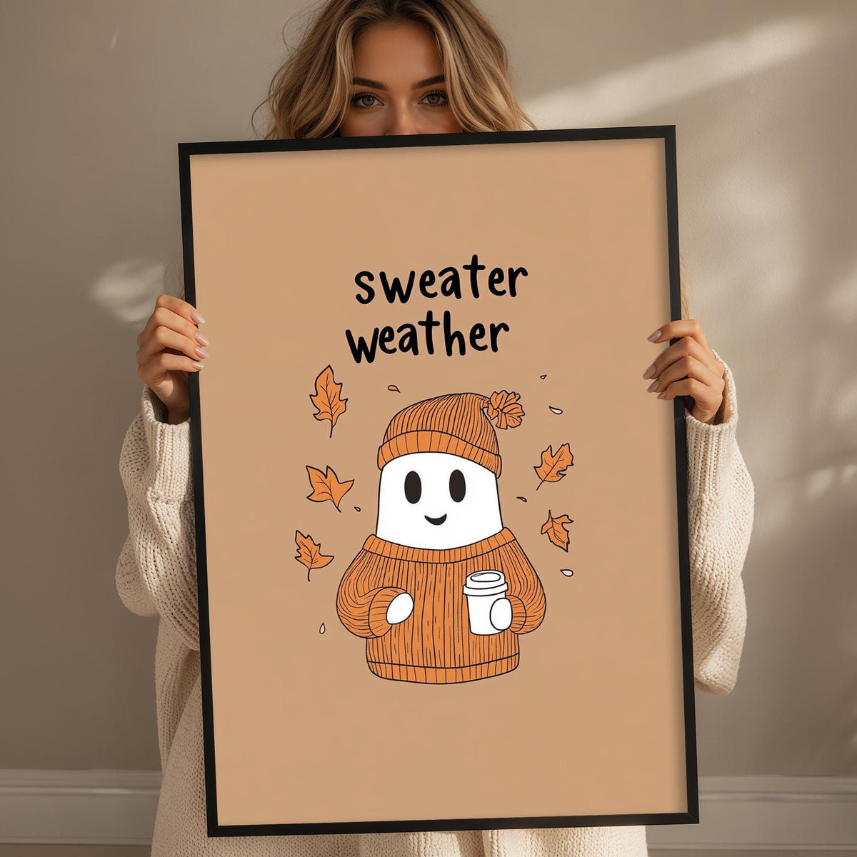 A woman holding a framed picture with a cartoon ghost wearing a sweater and holding a coffee cup, surrounded by autumn leaves.