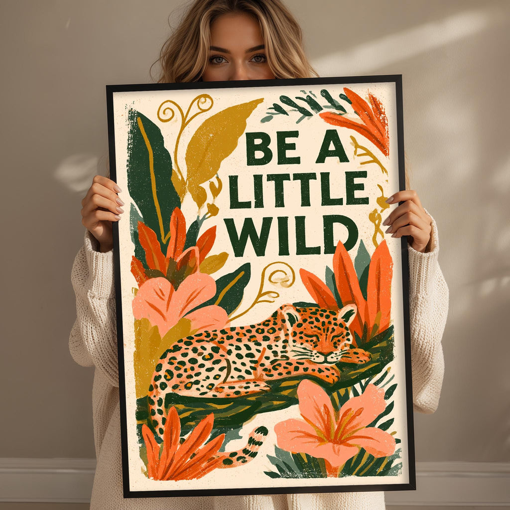 A woman is holding up a framed poster with a leopard and tropical flowers, with the text "Be a little wild" written on it.