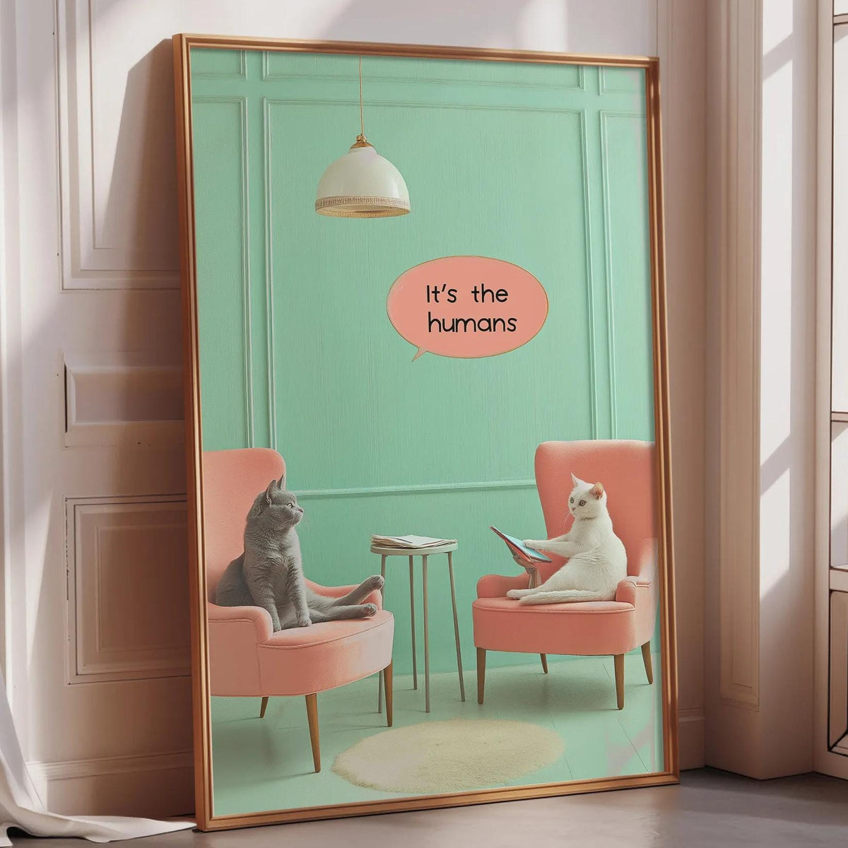 A framed poster depicting two cats sitting in chairs, with one cat appearing to read a book, and the other cat looking at the book. The text on the poster says "It's the humans".