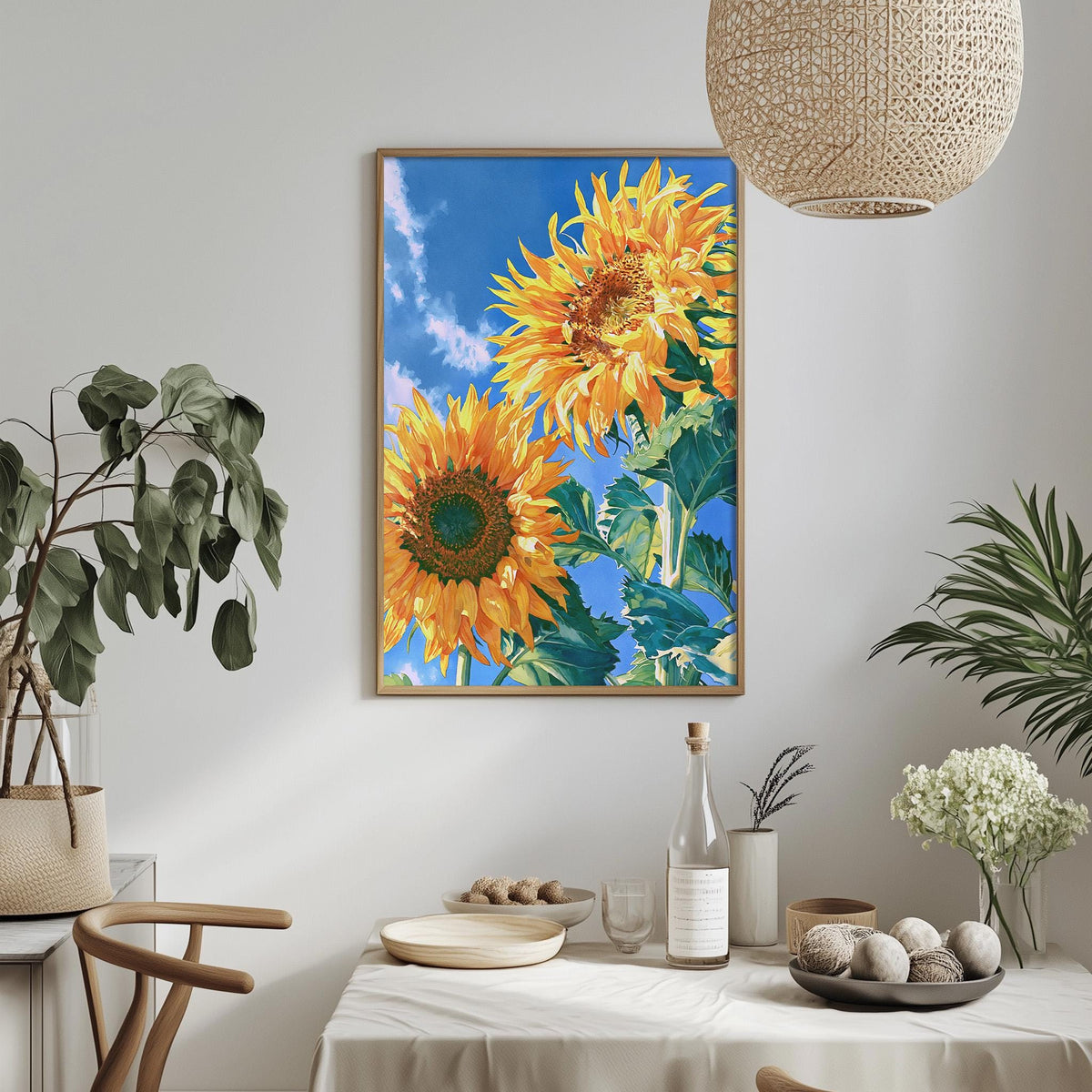 a dining table with a white tablecloth, a vase of white flowers, a bottle, and a bowl of fruit. Above the table, there is a framed painting of two sunflowers on a blue background.