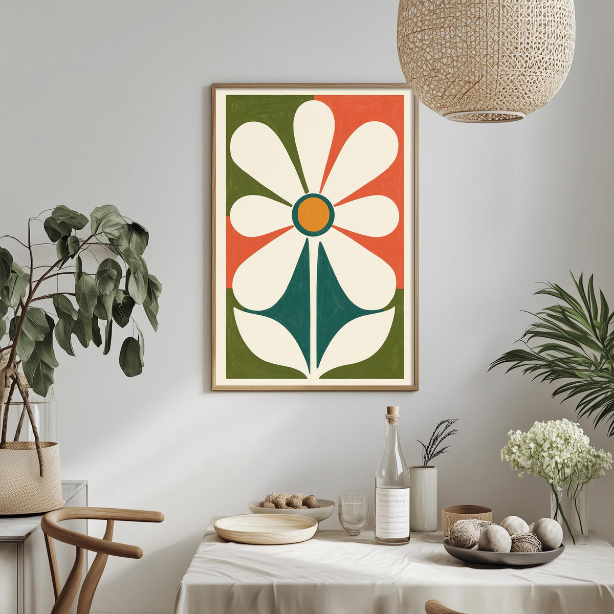 a dining table with a white tablecloth, a bowl of fruit, a bottle, and a vase with flowers. Above the table, there is a framed picture of a flower with a green stem and a white center. The table is set with a plate, a wine glass, and a bottle of wine. The room has a minimalist and modern design, with a woven light fixture hanging above the table and potted plants adding a touch of greenery.