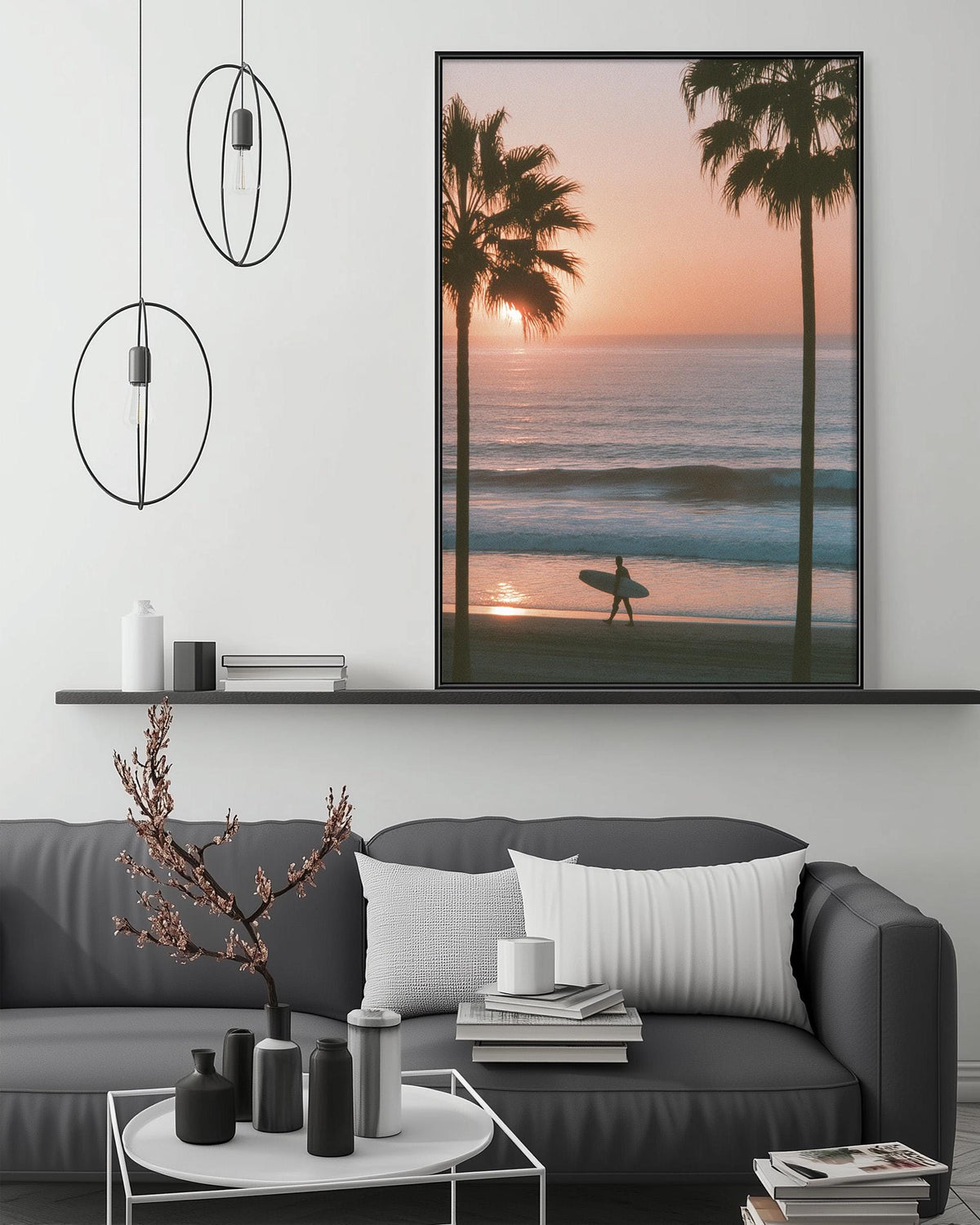 a serene beach scene with a surfer silhouetted against the setting sun, as depicted in a framed artwork hanging on the wall above a gray couch.