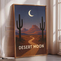 A framed poster depicting a desert landscape with a cactus in the foreground and a crescent moon in the night sky, with the text "DESERT MOON" below the cactus.