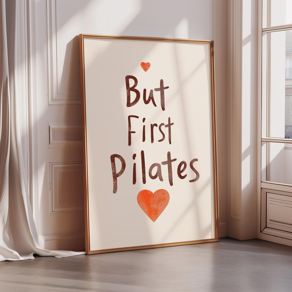 A framed poster with the text "But First Pilates" and an orange heart symbol is leaning against a white wall.
