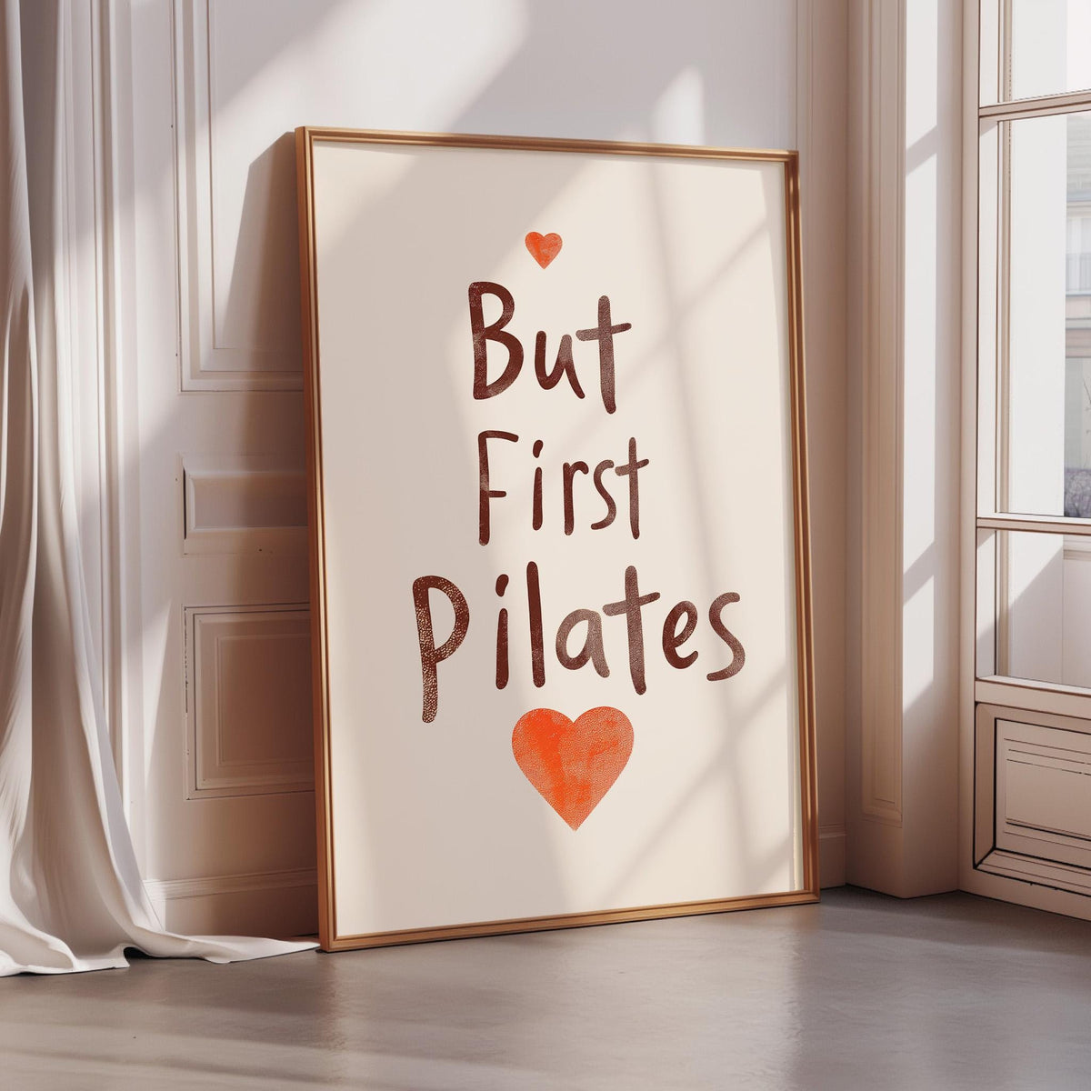 A framed poster with the text "But First Pilates" and an orange heart symbol is leaning against a white wall.
