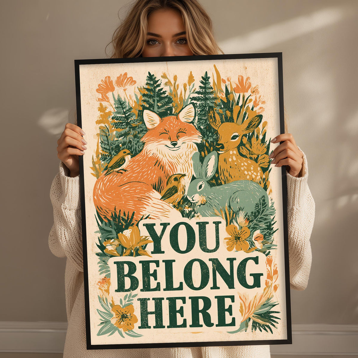 A woman is holding up a framed poster with a vibrant illustration of various animals, including a fox, rabbit, and deer, surrounded by lush foliage. The poster prominently displays the text "YOU BELONG HERE".