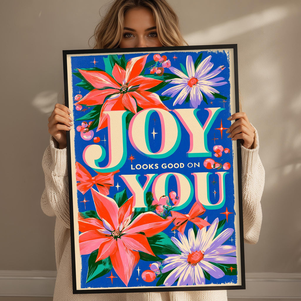 A woman is holding up a framed poster with a colorful floral design and the text "JOY LOOKS GOOD ON YOU".