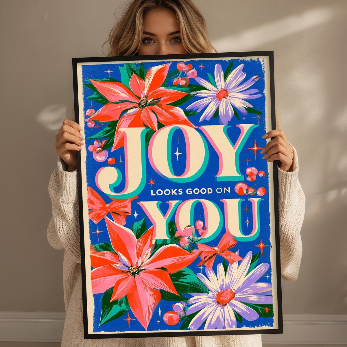 A woman is holding up a framed poster with a colorful floral design and the text "JOY LOOKS GOOD ON YOU".
