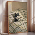 A framed painting depicting a black and white cat sitting on a striped blanket, holding a cup of coffee.