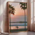 A framed photograph depicting a surfer walking along a beach at sunset, with palm trees in the foreground.
