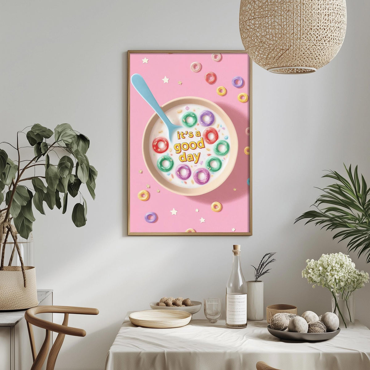 a framed poster with a colorful cereal bowl design, surrounded by a pink background and decorated with various donuts. The poster also displays the text "It's a good day" in a playful font. The poster is hung on a wall above a dining table, which has a vase of flowers, a bottle, and a bowl of fruit on it.