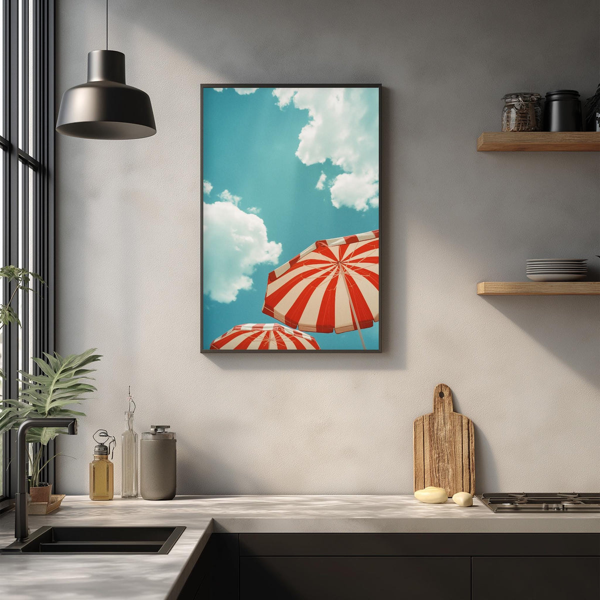 a kitchen counter with a framed picture of two red and white striped umbrellas against a blue sky with white clouds. The kitchen counter also has various kitchen items such as a cutting board, a bottle, and a bowl.