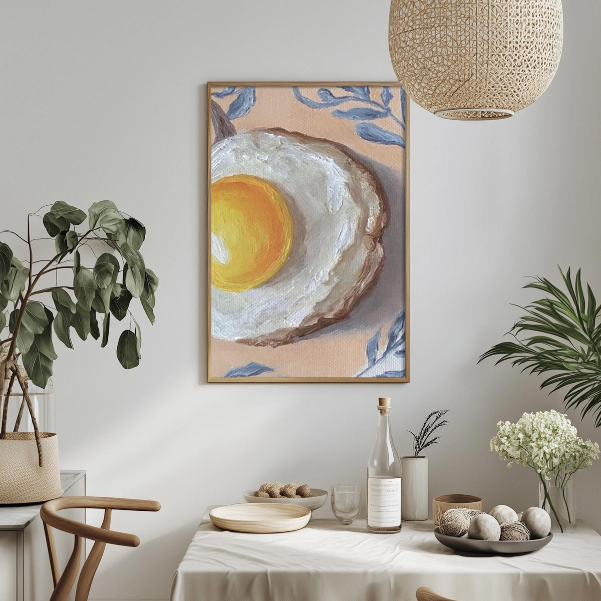 a dining table with a white tablecloth, a bowl of fruit, a bottle, and a vase with flowers. Above the table, there is a framed painting of an egg on a white background.