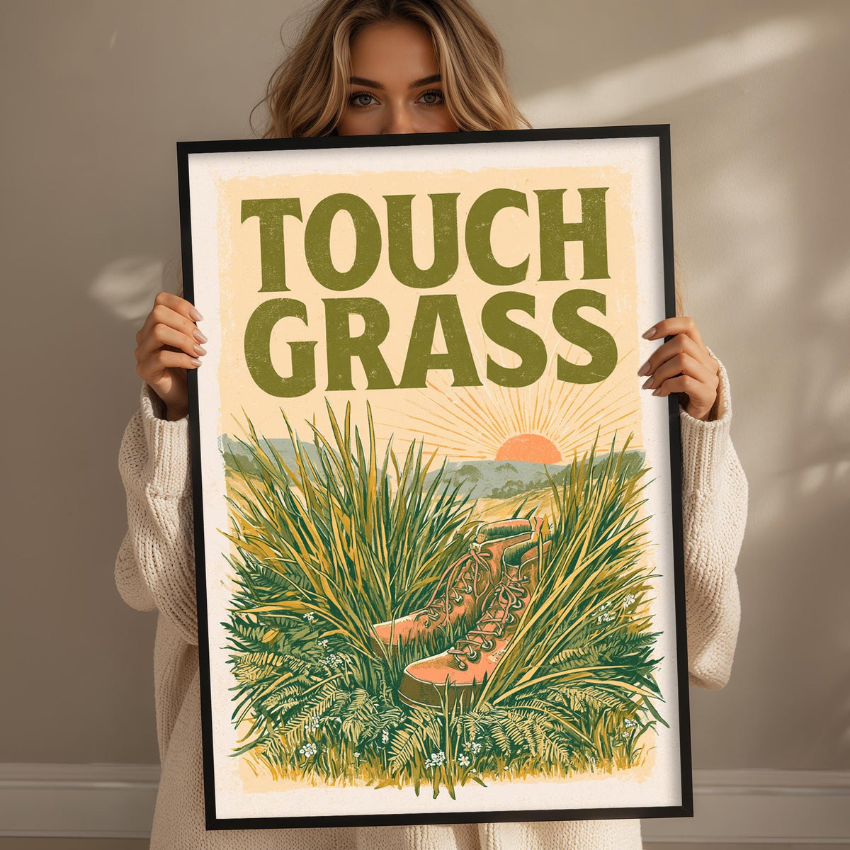 A woman holding a framed poster with a picture of a pair of hiking boots in tall grass, with the text "TOUCH GRASS" prominently displayed.