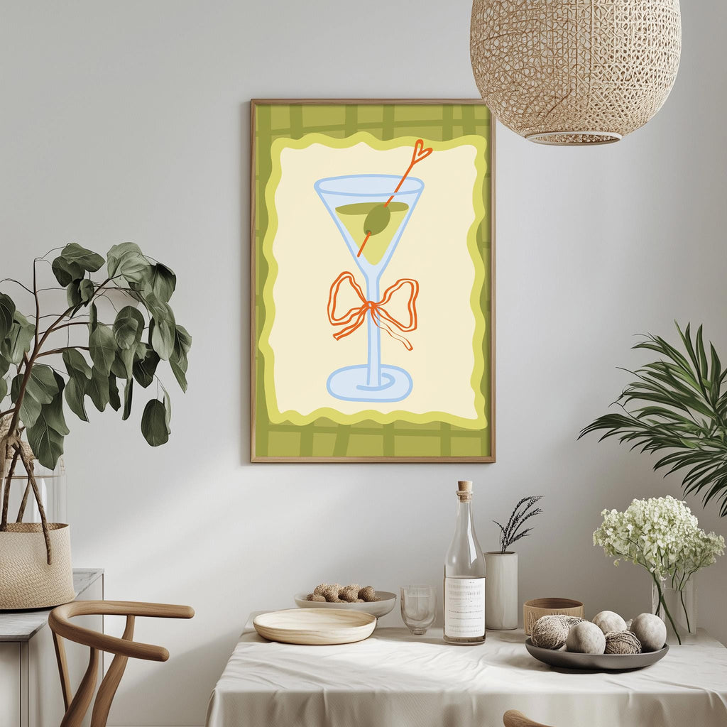 a dining table with a white tablecloth, a bowl of fruit, a bottle, and a vase with flowers. Above the table, there is a framed picture of a martini glass with a bow.