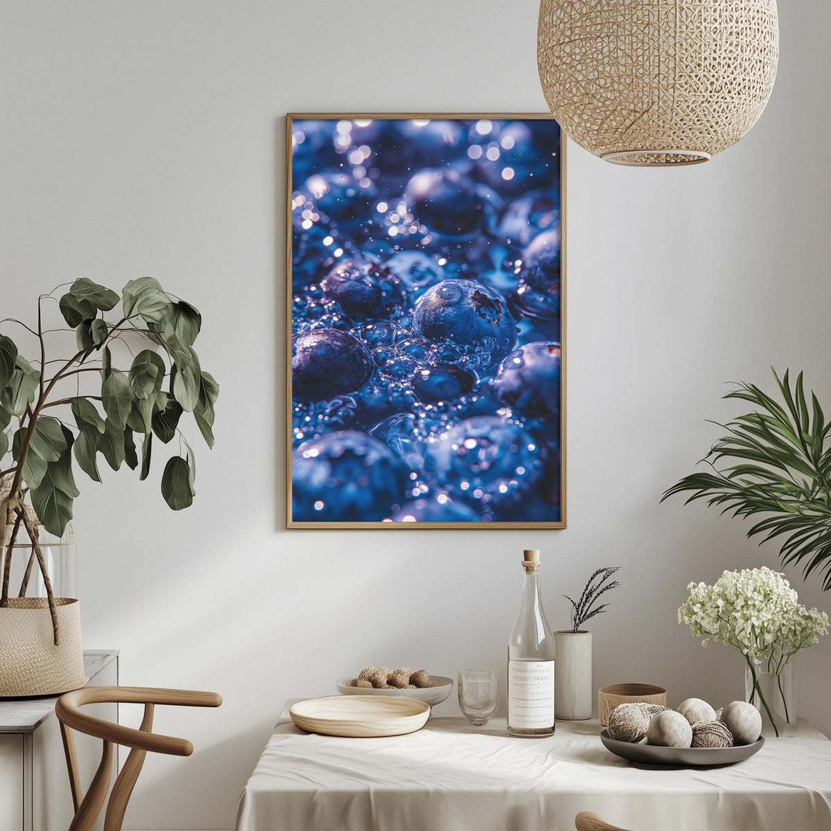 a dining table with a white tablecloth, a bowl of fruit, a bottle, and a vase with flowers. A framed picture of blueberries hangs on the wall above the table.