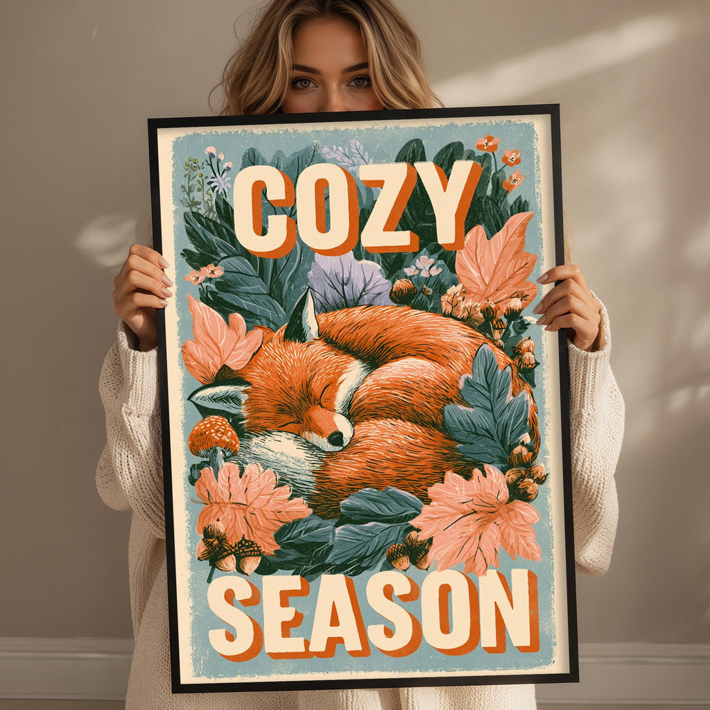 A woman is holding up a framed poster with a sleeping fox surrounded by flowers and leaves, with the text "COZY SEASON" written on it.