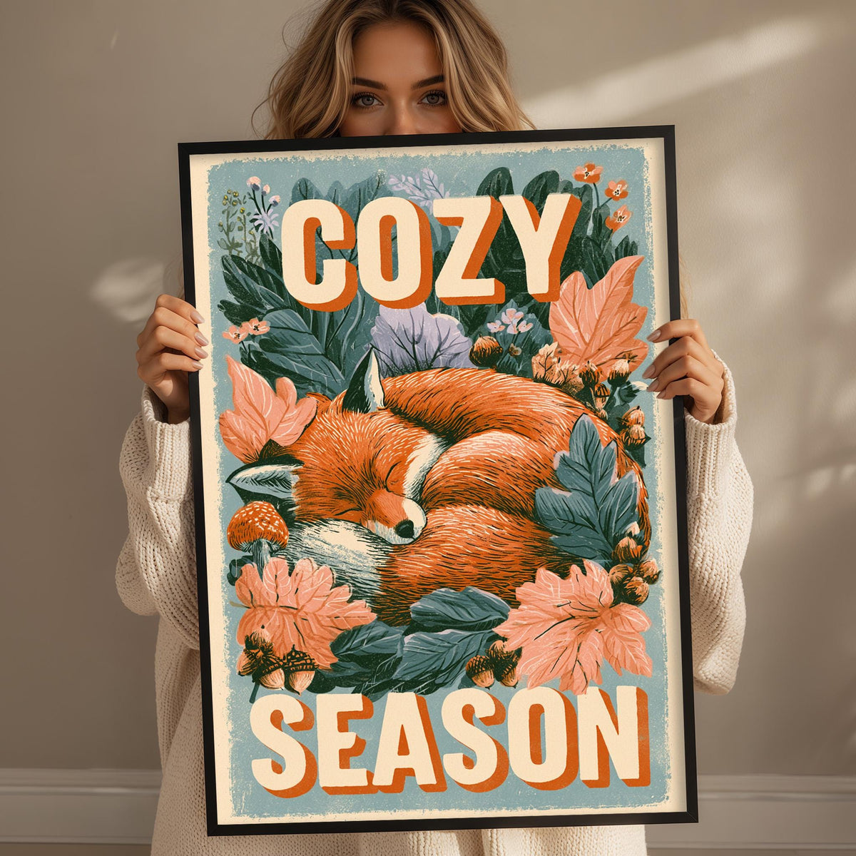 A woman is holding up a framed poster with a sleeping fox surrounded by flowers and leaves, with the text "COZY SEASON" written on it.