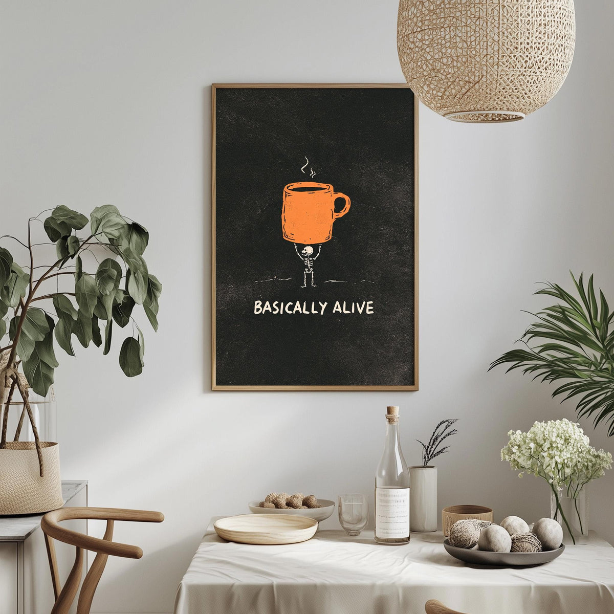 a dining table with a white tablecloth, a bowl of fruit, a bottle, and a vase with flowers. Above the table, there is a framed poster with an illustration of a coffee cup and the text "Basically alive".