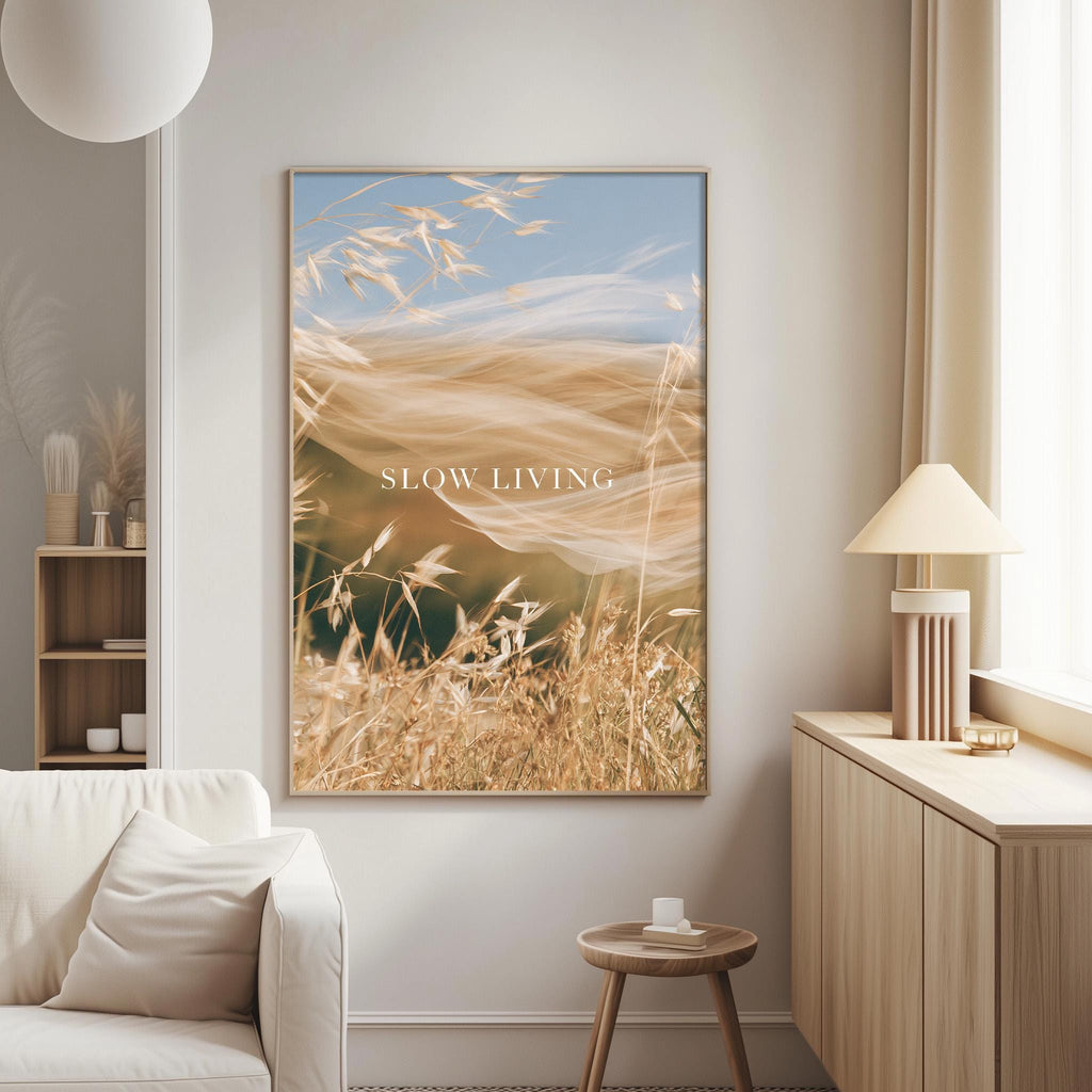 a living room with a large framed picture of a field of tall grass hanging on the wall. The picture has a serene and peaceful atmosphere, with the grass swaying gently in the breeze. The text "SLOW LIVING" is displayed on the picture, which adds a motivational and calming element to the room.