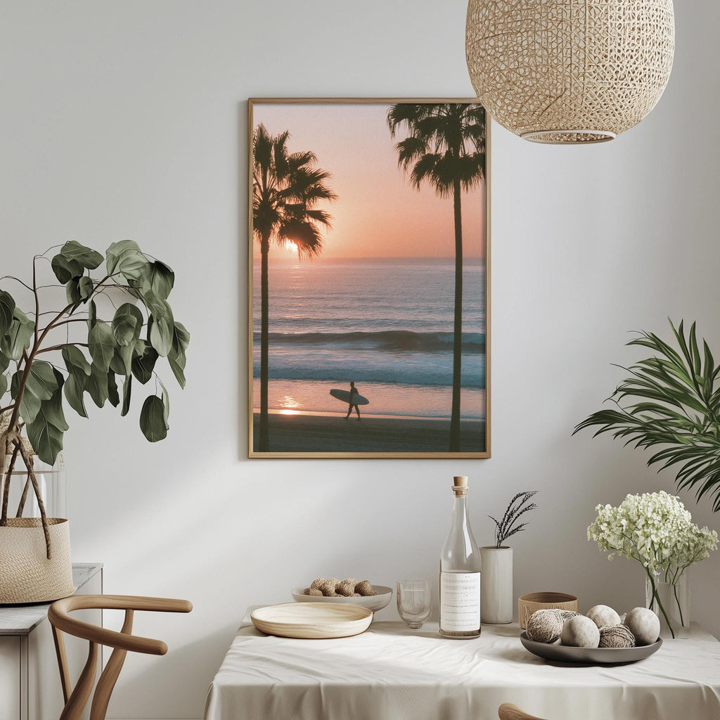 a serene beach scene with a surfer silhouetted against the setting sun, framed and hung on a wall. The table is set with a vase of flowers, a bottle, and a bowl of fruit, creating a cozy and inviting atmosphere.