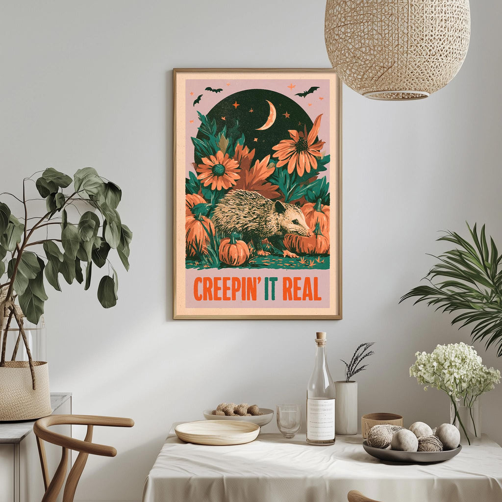 a framed poster with a whimsical illustration of a hedgehog surrounded by autumnal elements, including flowers and pumpkins, with the text "CREEPIN' IT REAL" displayed prominently. The poster is hung on a wall above a dining table, which has various items such as a bottle, a bowl, and a vase. The room also includes potted plants and a hanging light fixture, creating a cozy and inviting atmosphere.