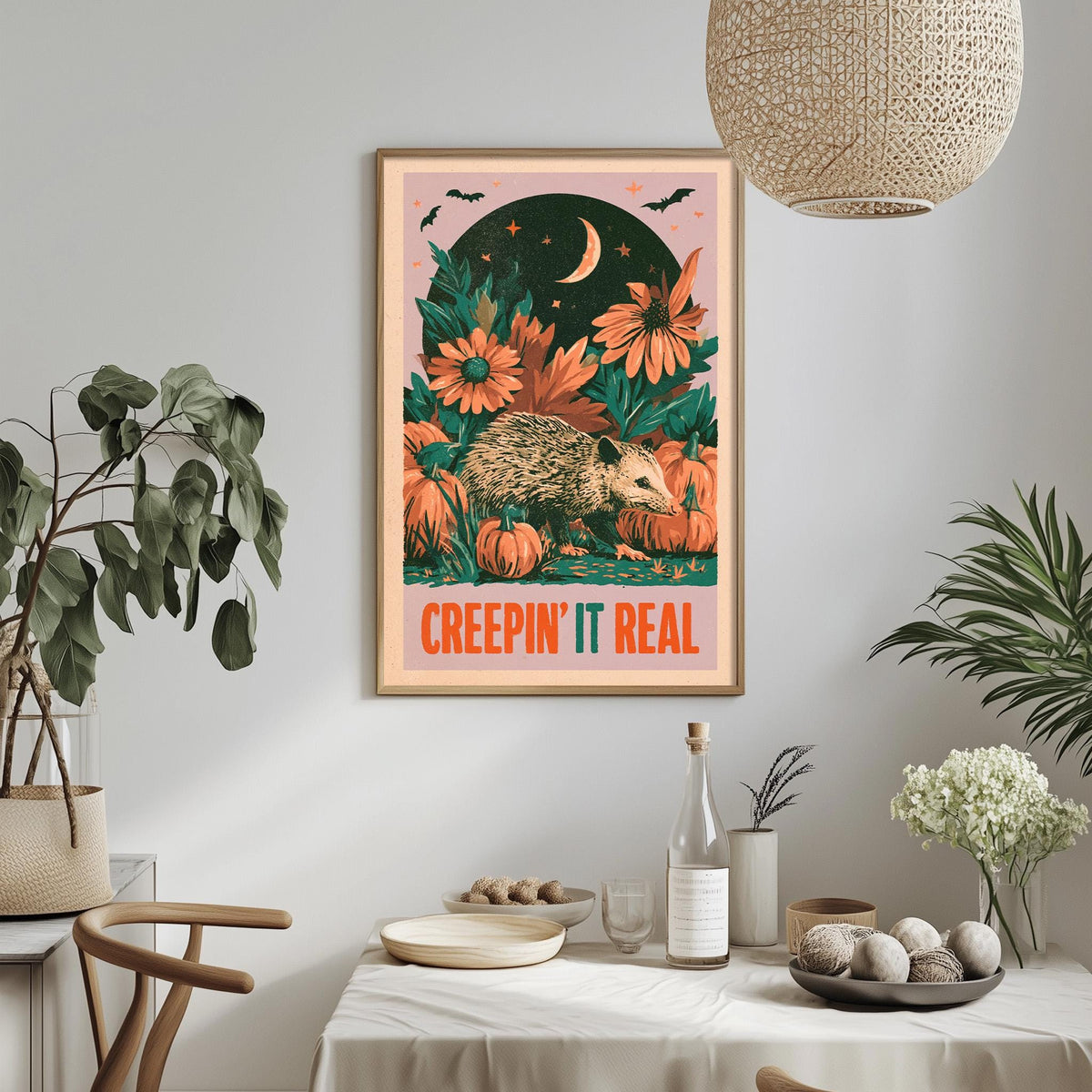 a framed poster with a whimsical illustration of a hedgehog surrounded by autumnal elements, including flowers and pumpkins, with the text "CREEPIN' IT REAL" displayed prominently. The poster is hung on a wall above a dining table, which has various items such as a bottle, a bowl, and a vase. The room also includes potted plants and a hanging light fixture, creating a cozy and inviting atmosphere.
