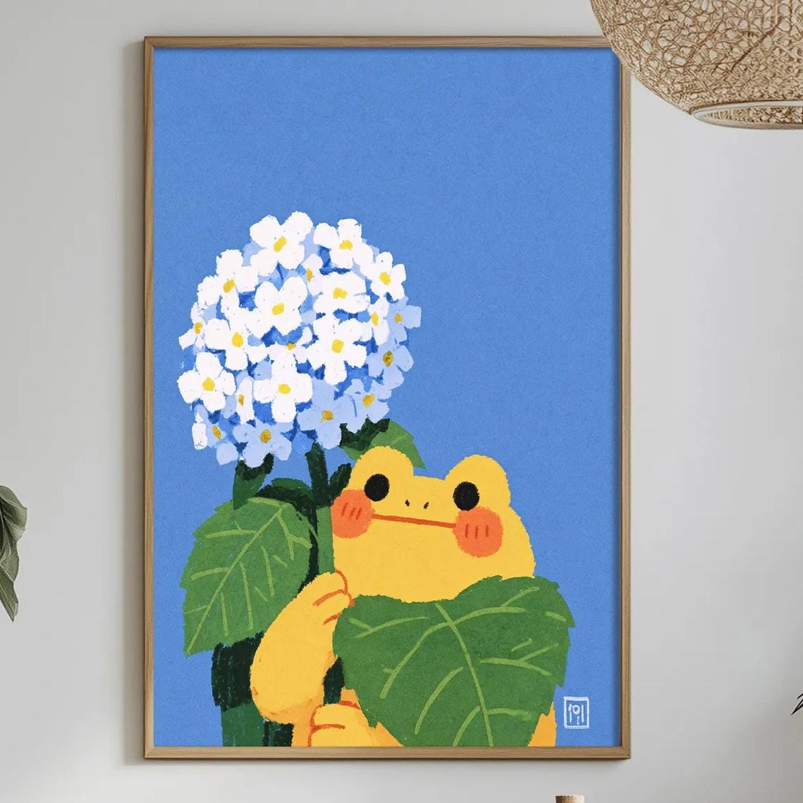 a framed illustration of a frog holding a bunch of white flowers, set against a blue background. The frog is surrounded by green leaves, and the flowers are blooming. The scene is set in a dining area with a table, chairs, and various decorative elements such as potted plants and vases.