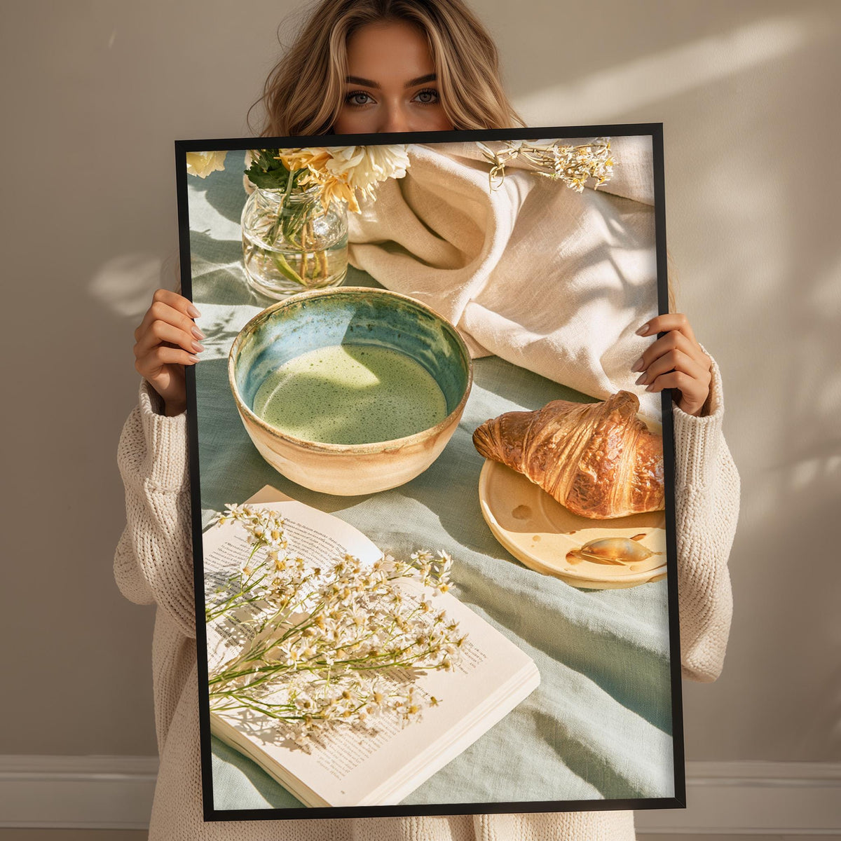 A woman holds up a framed image of a table with a croissant, a bowl of soup, and a book with flowers on the cover.