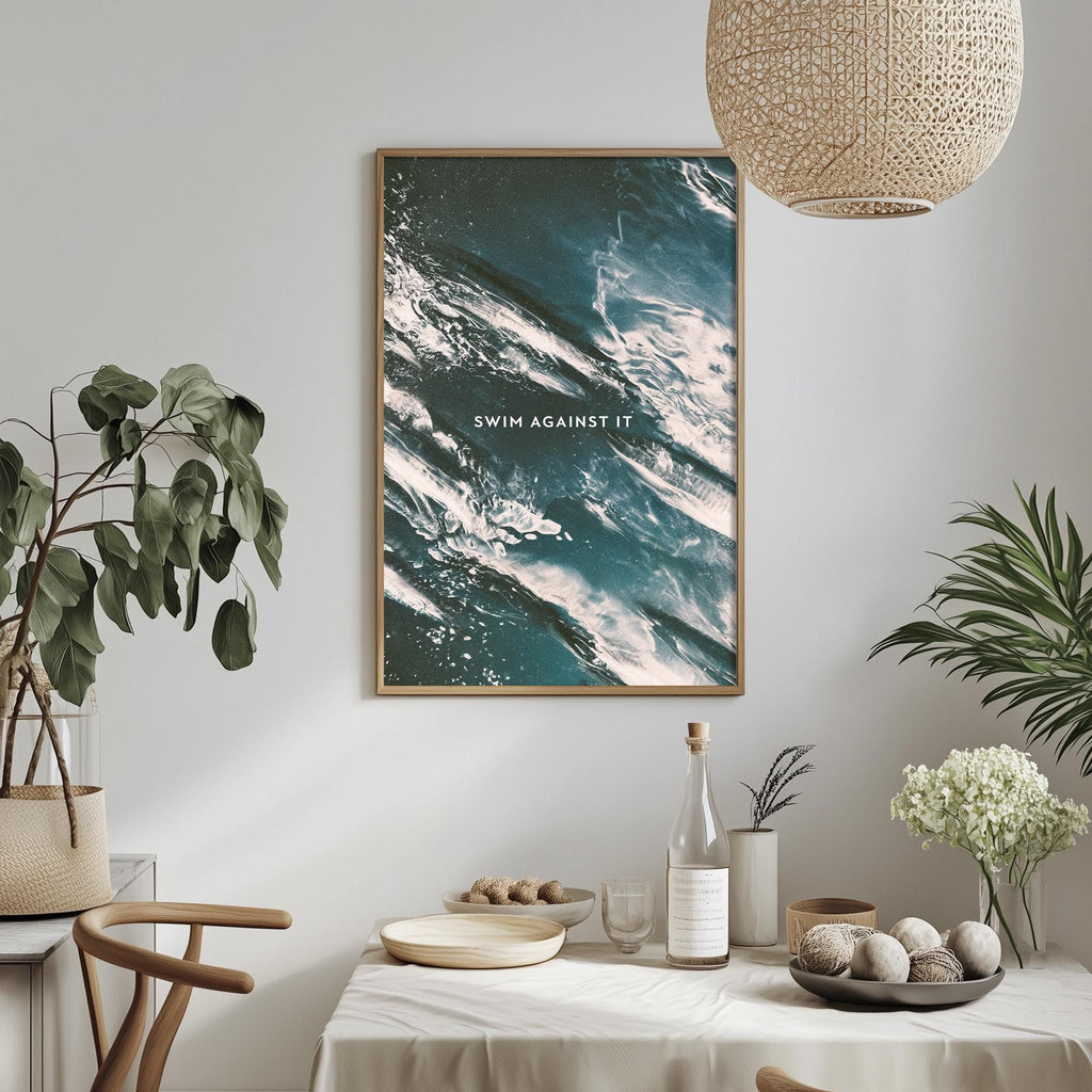 a dining table with a white tablecloth, a bowl of fruit, a bottle, and a vase with flowers. Above the table, there is a framed poster with the text "SWIM AGAINST IT" displayed on it.