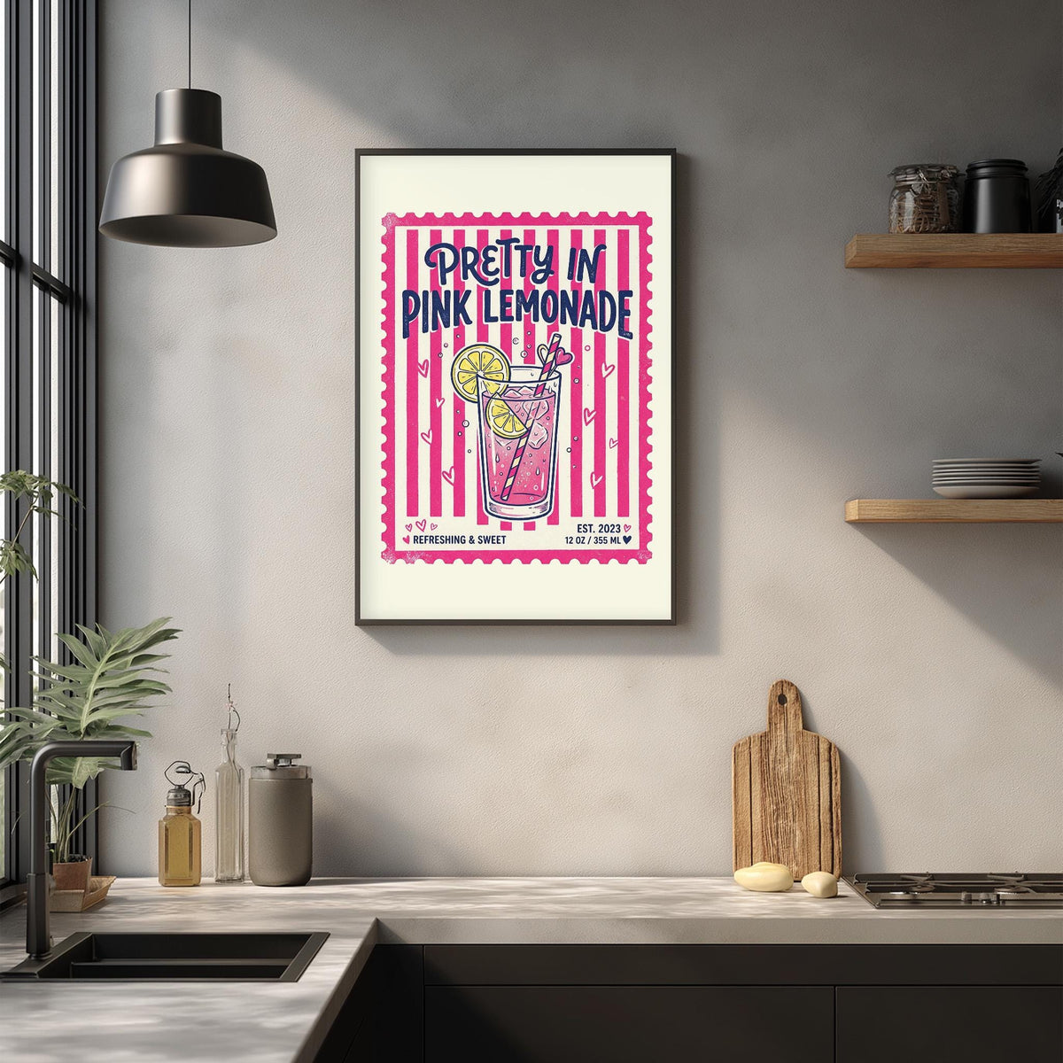 A framed poster of a pink lemonade drink hangs on a wall above a kitchen counter with various kitchen items and a potted plant.