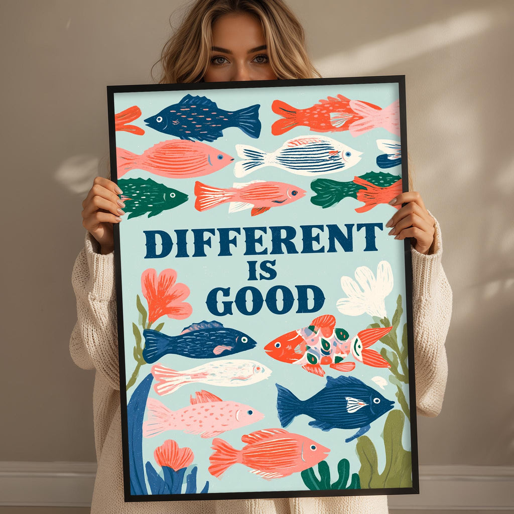 A woman is holding up a framed poster with a colorful illustration of fish and the text "DIFFERENT IS GOOD" written on it.