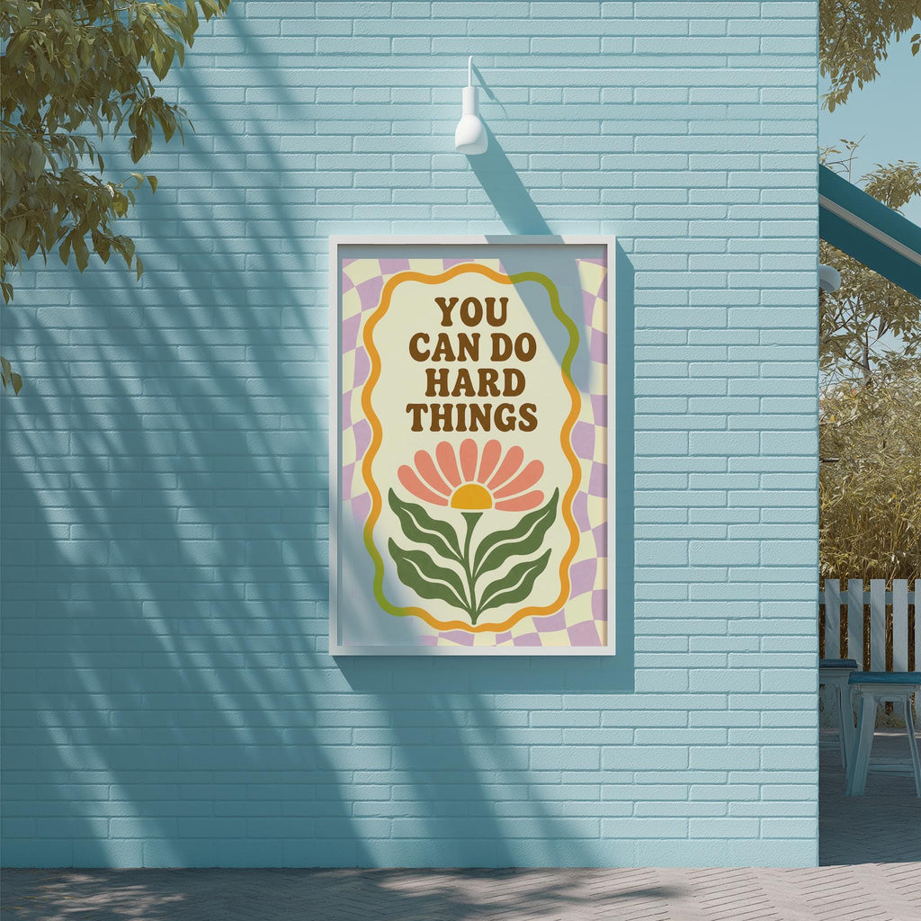 A framed poster with the text "YOU CAN DO HARD THINGS" is displayed on a blue brick wall, with a tree and a white light fixture visible in the background.
