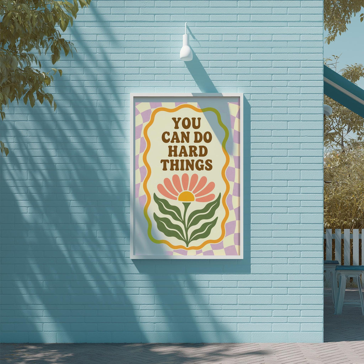 A framed poster with the text "YOU CAN DO HARD THINGS" is displayed on a blue brick wall, with a tree and a white light fixture visible in the background.
