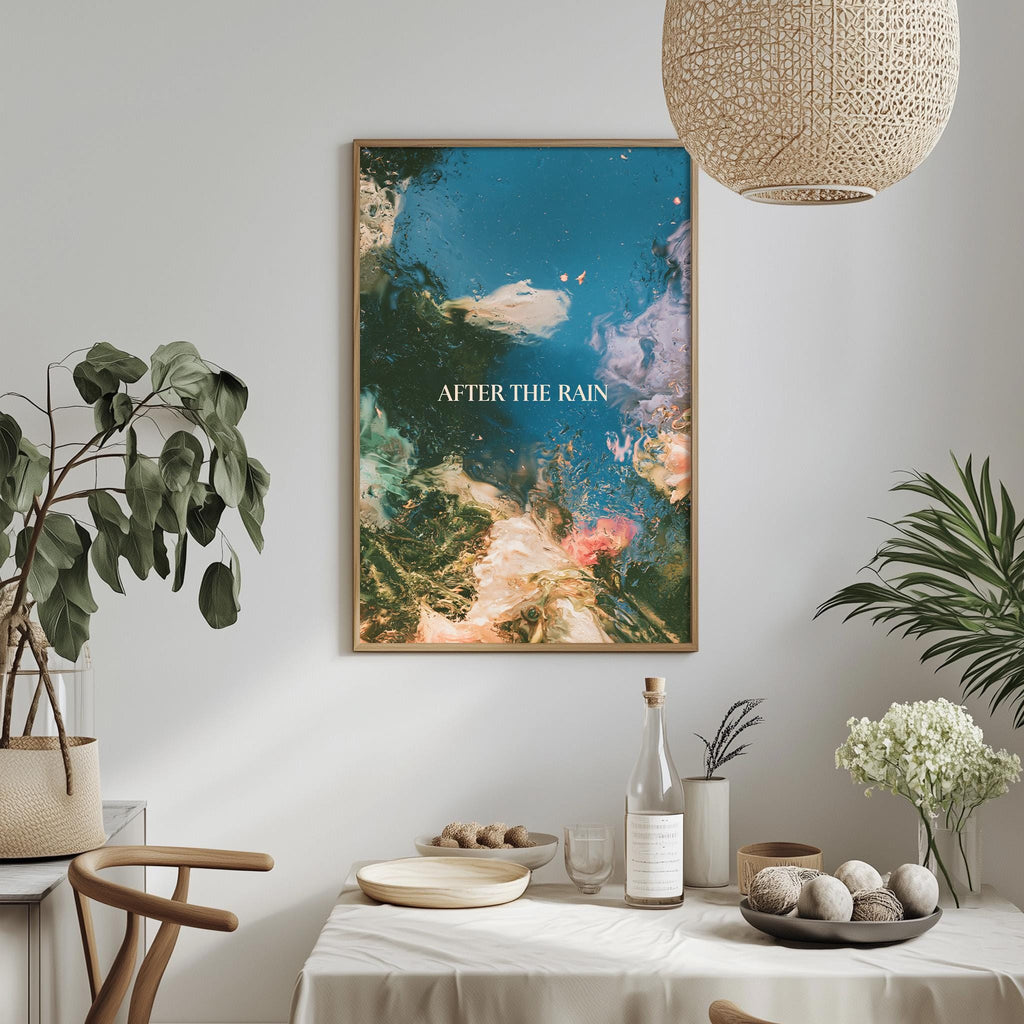 a dining table with a white tablecloth, a bowl of fruit, a bottle, and a vase with flowers. Above the table, there is a framed poster with the text "AFTER THE RAIN" displayed on it.