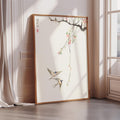 A framed painting depicting two birds in flight, with a branch with flowers and leaves in the background, is leaning against a wall in a room with sunlight streaming in through a window.