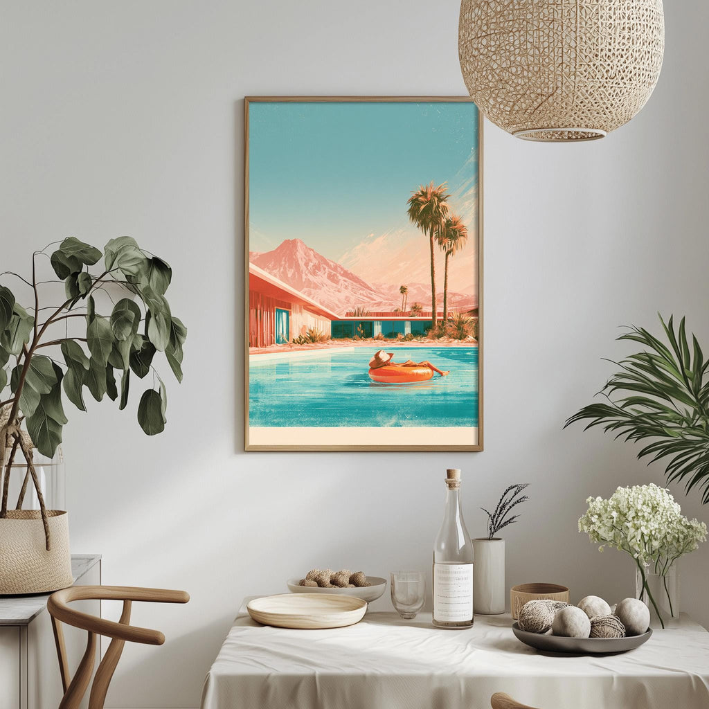 a framed painting of a person relaxing in a pool, surrounded by palm trees and mountains, with a beach house in the background. The painting is hung on a wall above a dining table with various decorative items and plants.