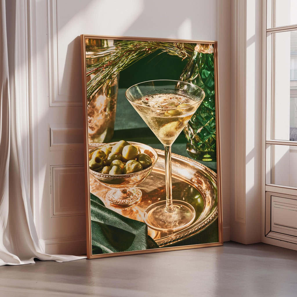 A framed photograph of a martini glass filled with a golden liquid, surrounded by a bowl of green olives and a bottle of olive oil, set against a white wall and a window.