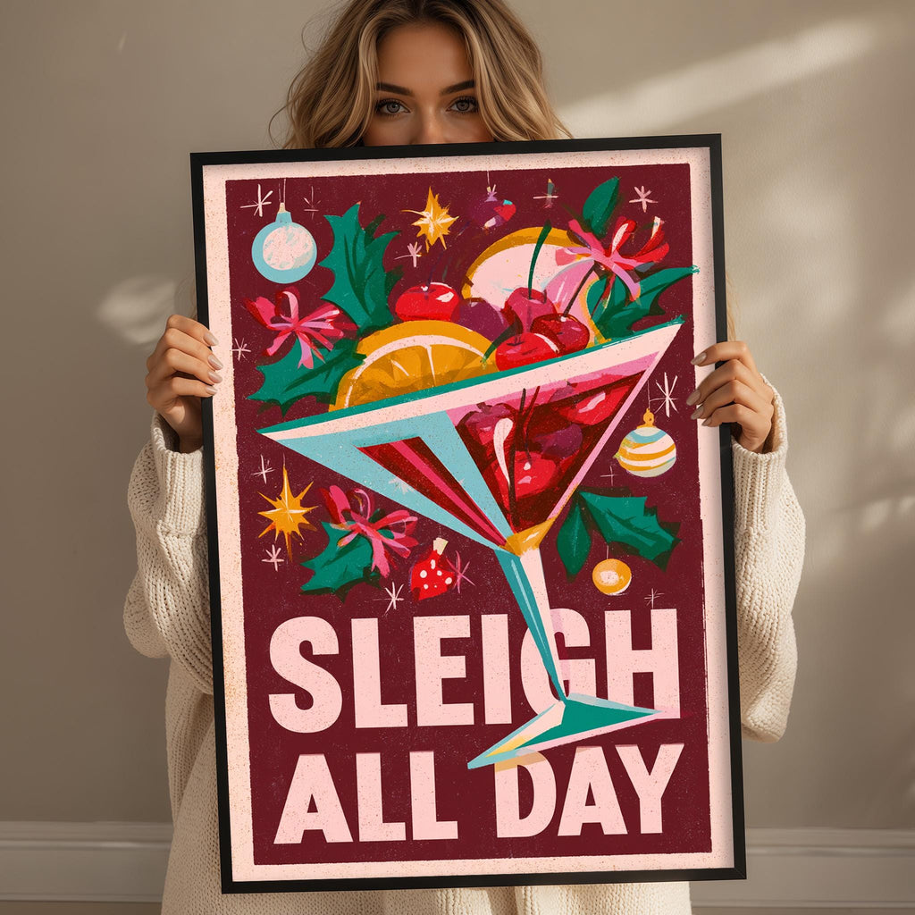 A woman is holding up a framed poster with a colorful illustration of a cocktail glass filled with fruit and flowers, along with the text "SLEIGH ALL DAY".