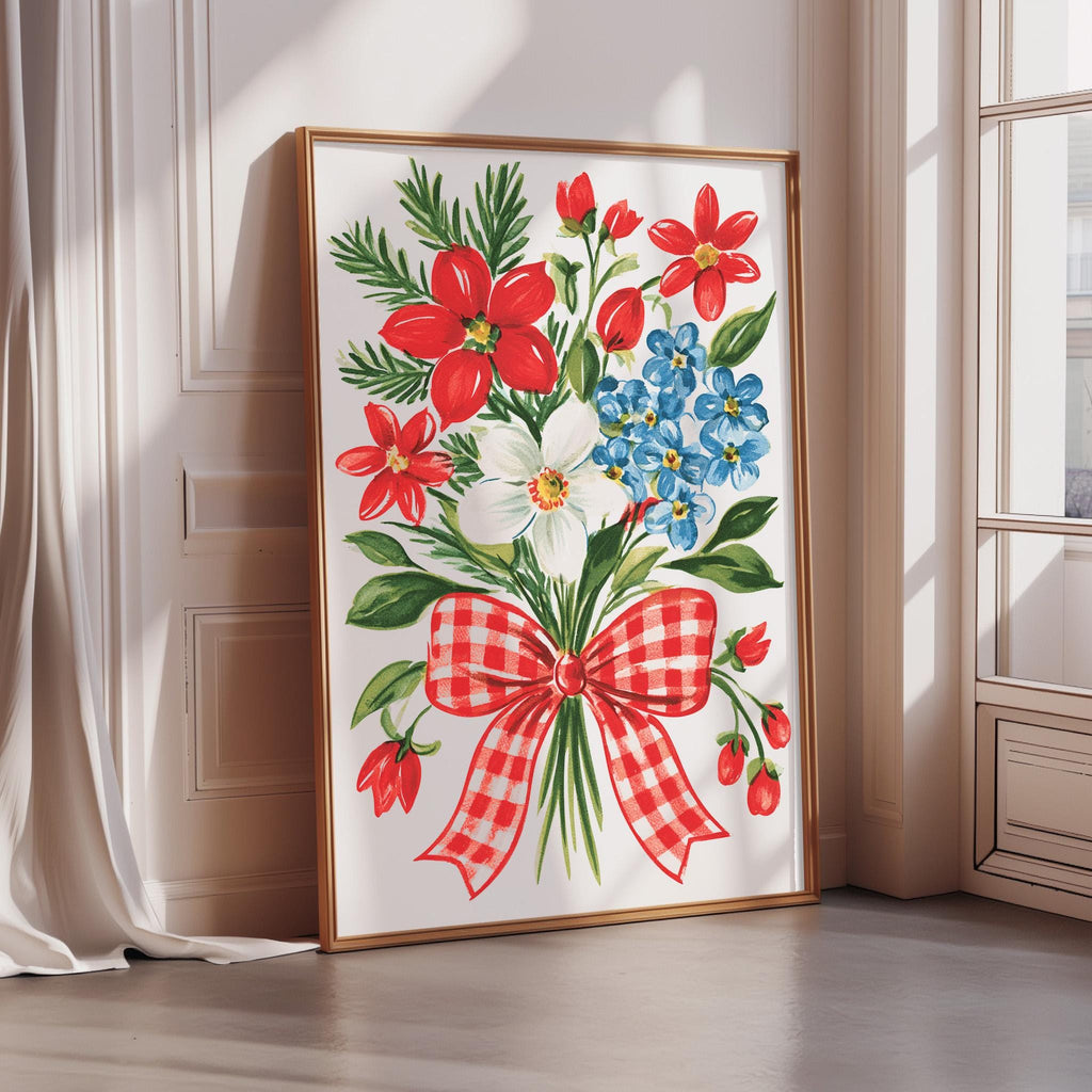 A framed painting depicting a vibrant bouquet of red, white, and blue flowers, with a red and white checkered bow, is displayed on a wall in a room with a window.