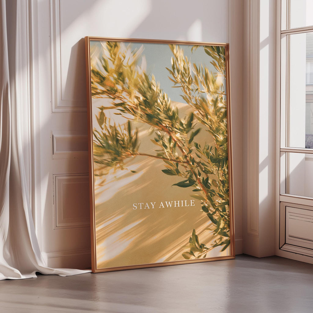 A framed poster with a photograph of a tree and the text "STAY AWHILE" is displayed on a wall in a room with sunlight streaming in through a window.