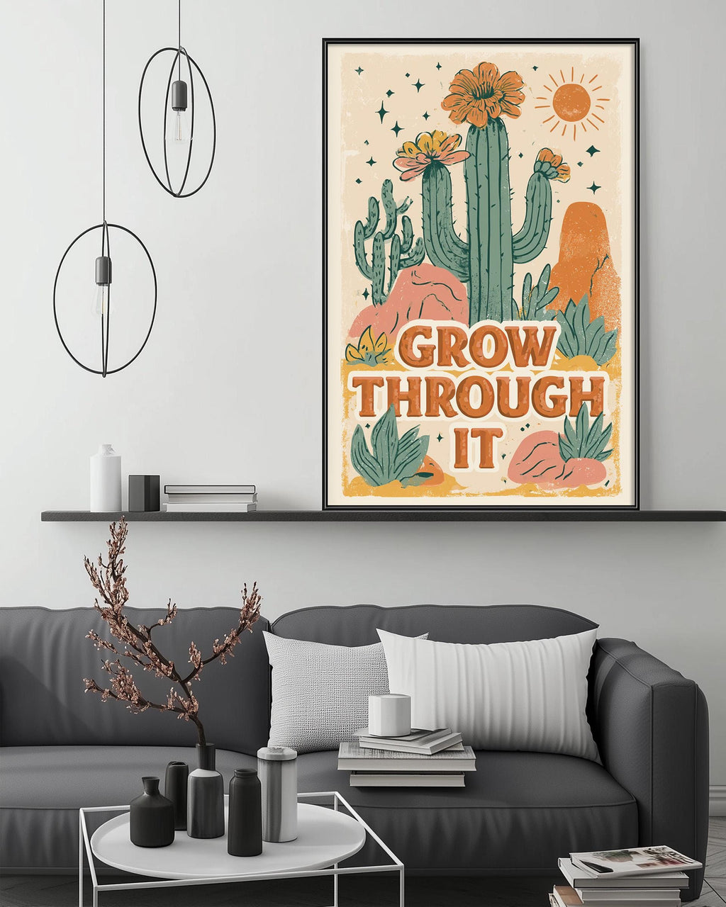 a framed poster with a desert-themed illustration, including cacti, flowers, and a sun, hanging on a wall above a gray couch with a coffee table in front of it.