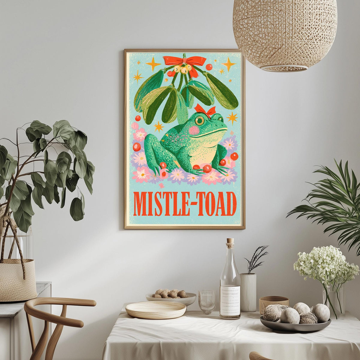 a framed poster of a green frog sitting under a mistletoe, surrounded by flowers and stars, hanging on a wall above a dining table with various items on it.